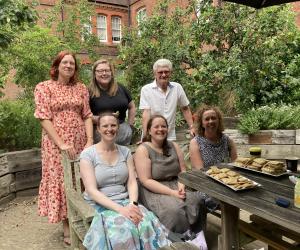 Image of volunteers and staff at a small garden party celebrating the first 100 transcriptions. Its shows people sitting by a table which has trays of biscuites on it
